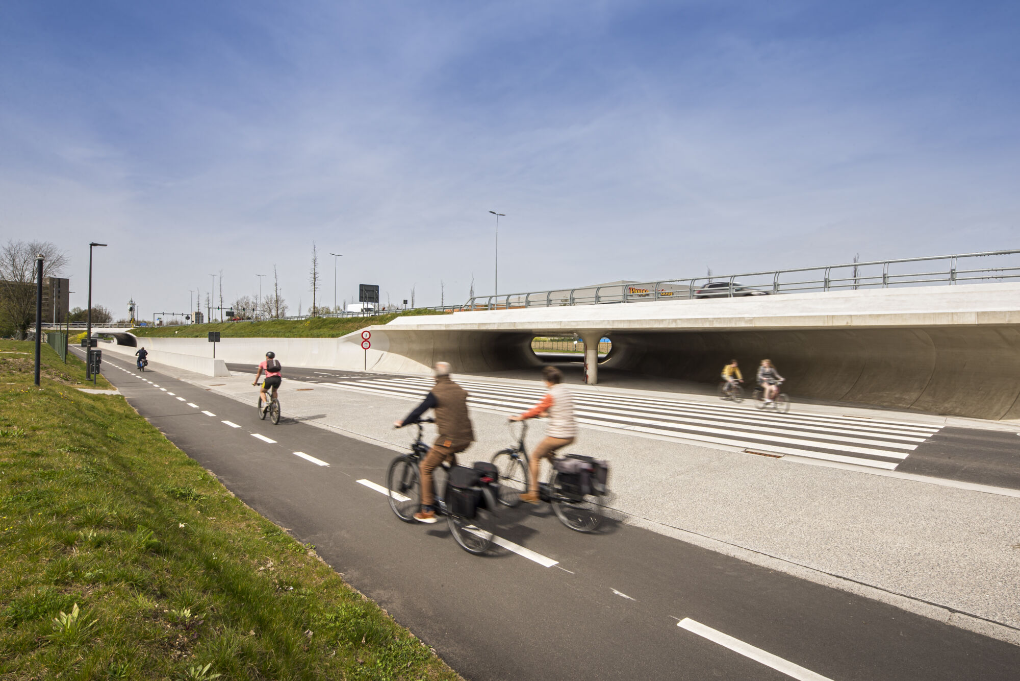 Fietsers fietsen op een tweerichtingsfietspad, afgescheiden van het autoverkeer. In de verte zie je nog fietsers in een nieuwe fietserstunnel.